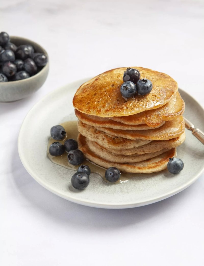 A stack of American-style pancakes topped with blueberries and drizzled with syrup sits on a plate, reminiscent of a mouthwatering recipe from the Hairy Bikers' Chicken &amp; Egg series. A bowl of fresh blueberries awaits beside it.
