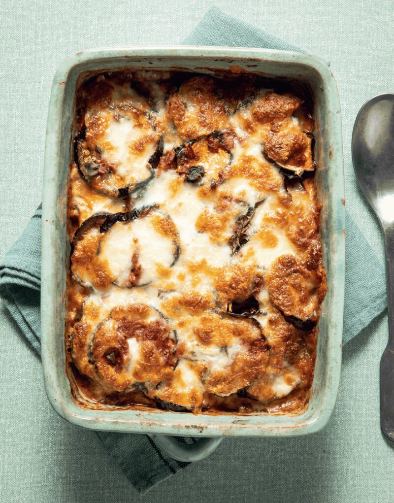A baked casserole dish of golden-brown, cheesy Aubergine Parmigiana inspired by the Hairy Dieters’ Simple Healthy Food sits on a folded green cloth next to a metal spoon, viewed from above.