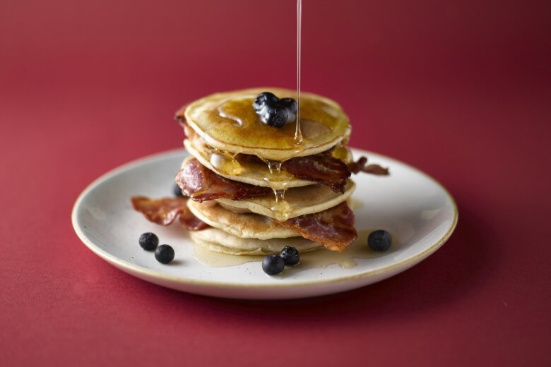 A stack of pancakes topped with blueberries and crispy bacon, drizzled with syrup, served on a white plate against a red background, reminiscent of the recipes in Hairy Bikers 'Mum's Know Best' cookery book.