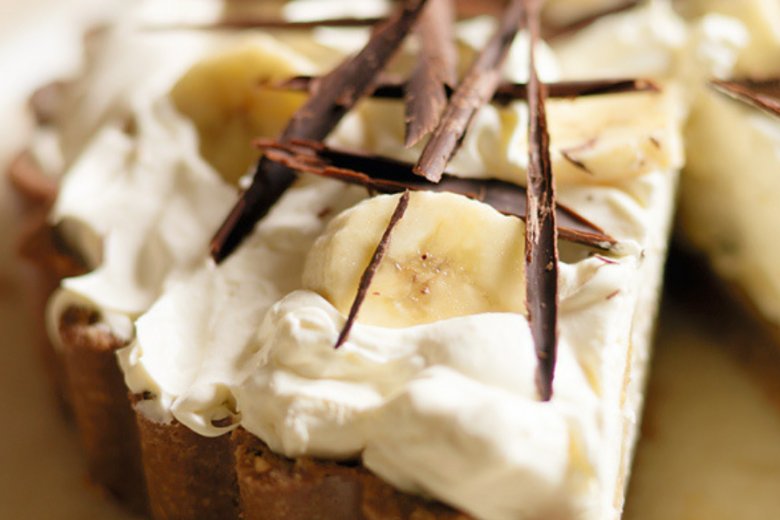 A close-up of a banana cream pie inspired by the Hairy Bikers, topped with whipped cream, banana slices, and decorative shards of dark chocolate, with one slice partially removed.
