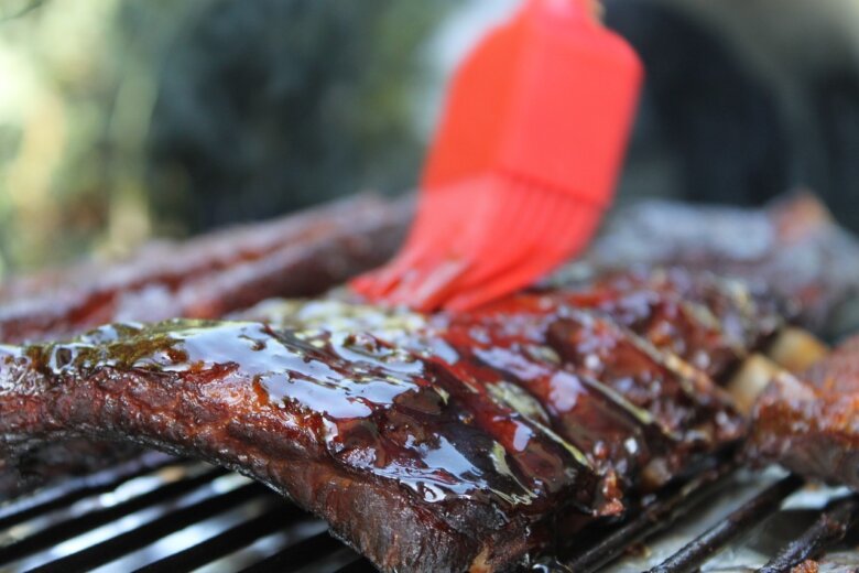A red brush applies sauce to a set of delicious BBQ ribs on a grill.