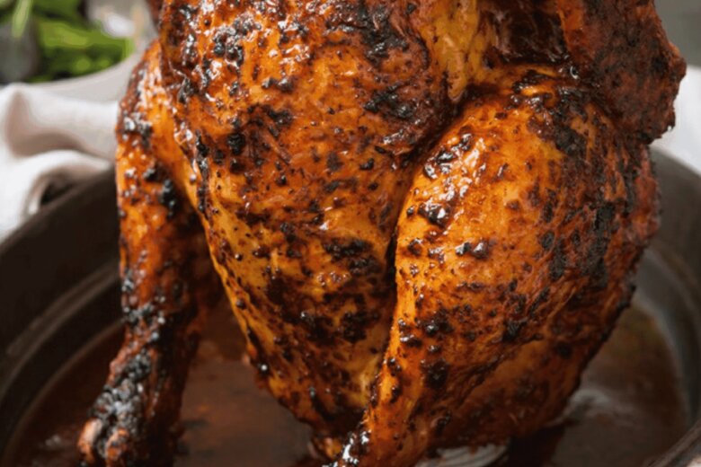 Whole roasted chicken with a crispy, seasoned skin, standing upright in a black roasting pan, resembling the mouthwatering Beer Can Chicken from a Hairy Bikers recipe, with herbs in the background.