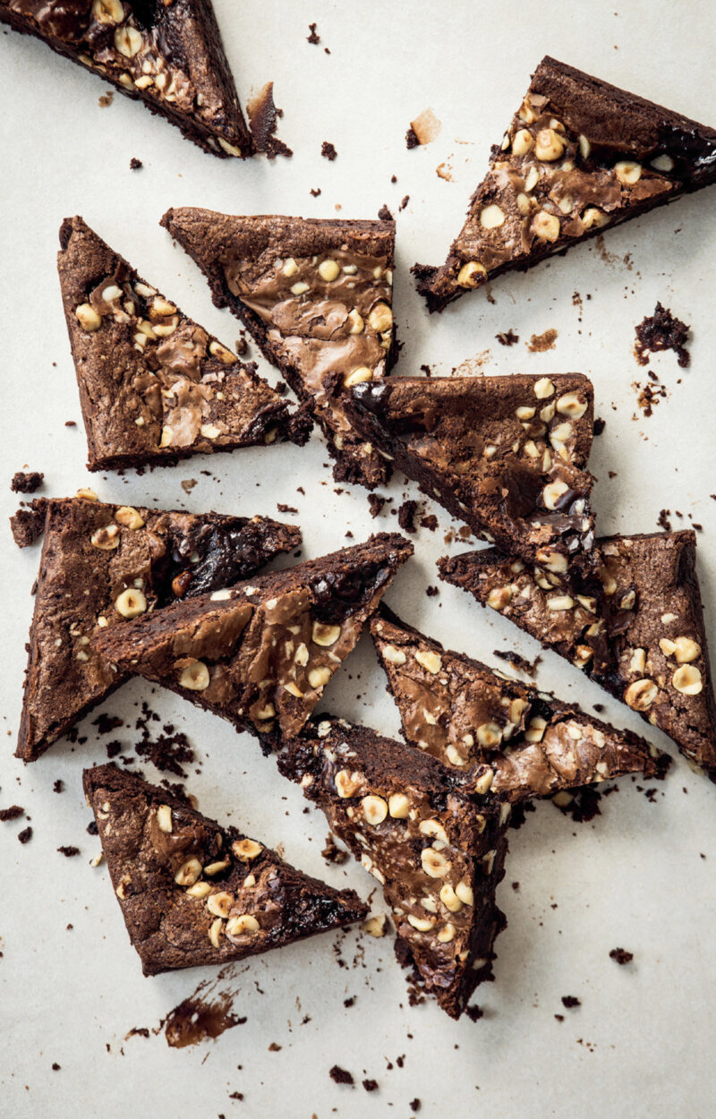 Sliced triangles of Biker Brownies with hazelnuts are scattered on a light surface, showing their rich, fudgy texture and nut pieces throughout. Crumbs are visible around the brownies, inspired by a Hairy Bikers recipe.