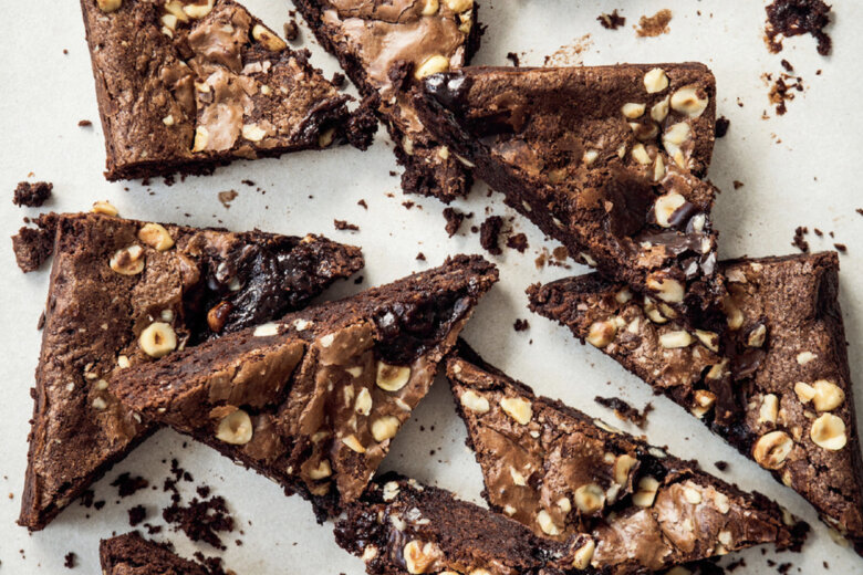 Sliced triangles of Biker Brownies with hazelnuts are scattered on a light surface, showing their rich, fudgy texture and nut pieces throughout. Crumbs are visible around the brownies, inspired by a Hairy Bikers recipe.