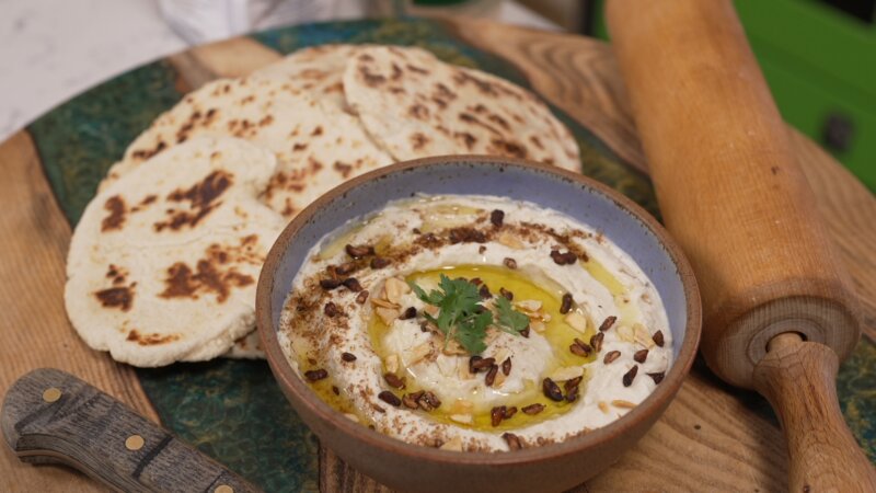 A bowl of butterbean hummus garnished with herbs, nuts, and olive oil, placed on a green plate with several pieces of grilled flatbreads and a rolling pin beside it.