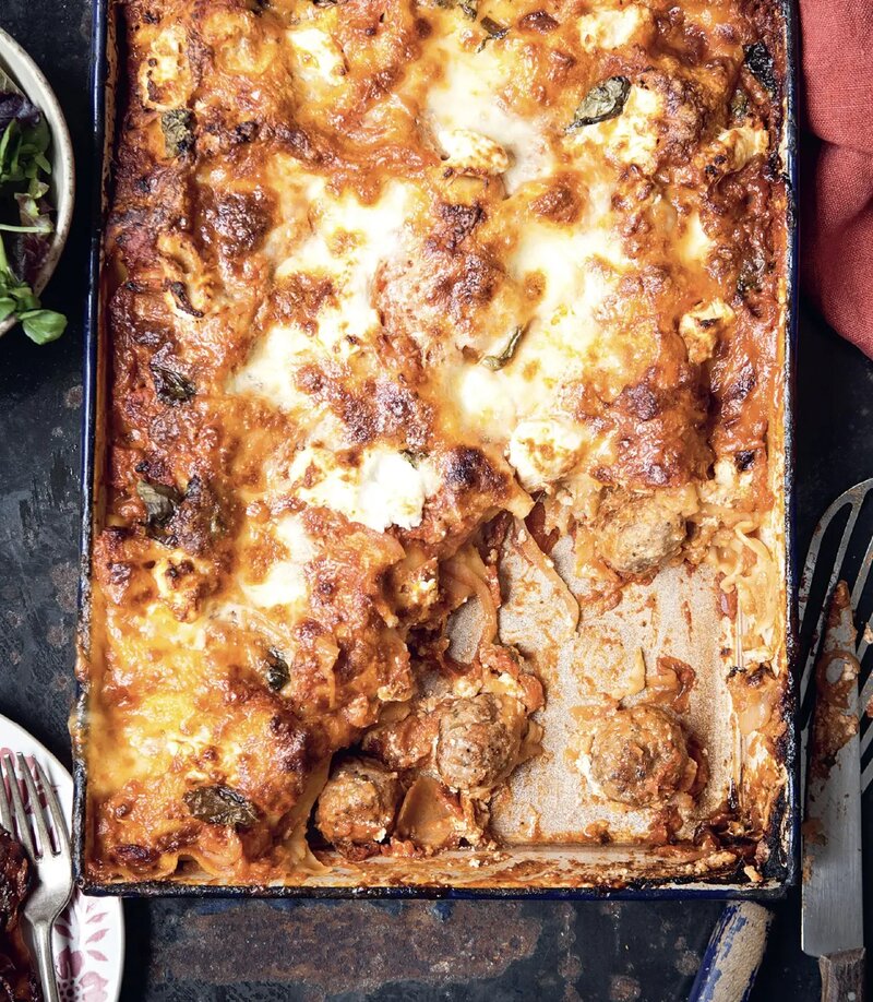 A celebratory lasagne sits in a rectangular pan with some portions removed, revealing layers of pasta, meatballs, sauce, and cheese. A plate with salad and cutlery rests beside the pan, reminiscent of the comfort found in the best of the Hairy Bikers' creations.
