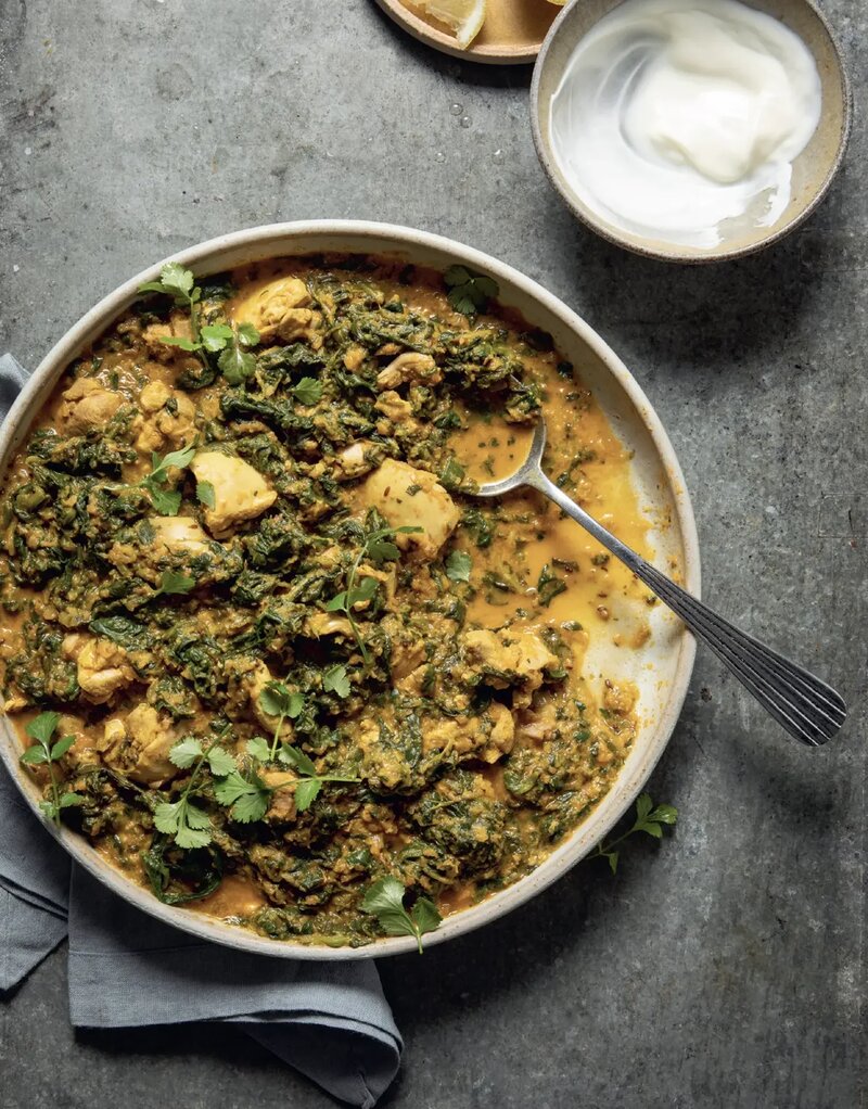 A bowl of Chicken &amp; Spinach Curry, with tender chicken and spinach in a creamy sauce, is garnished with coriander. Inspired by Hairy Dieters Simple Healthy Food, it’s served with yoghurt on the side atop a grey surface.