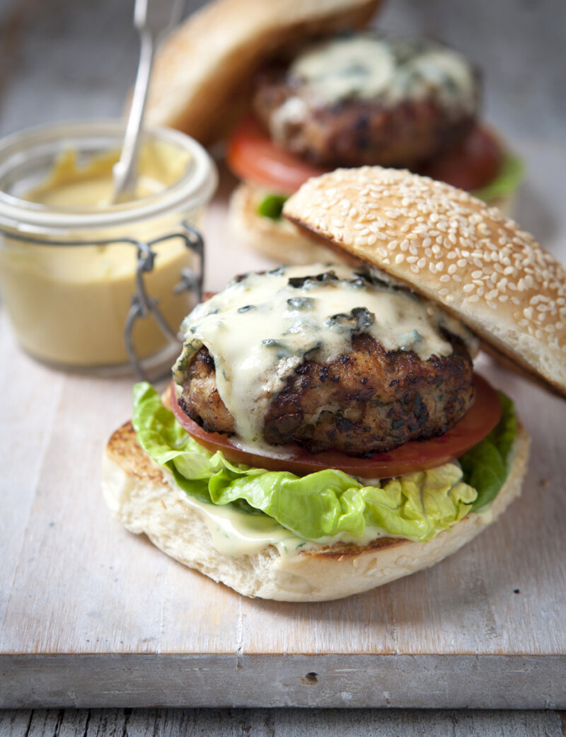 A close-up of a chicken burger from the Hairy Bikers 'chicken and egg' cookbook, with a sesame seed bun, lettuce, tomato, grilled patty topped with melted blue cheese, and a jar of creamy sauce in the background. Another burger is slightly out of focus behind it.