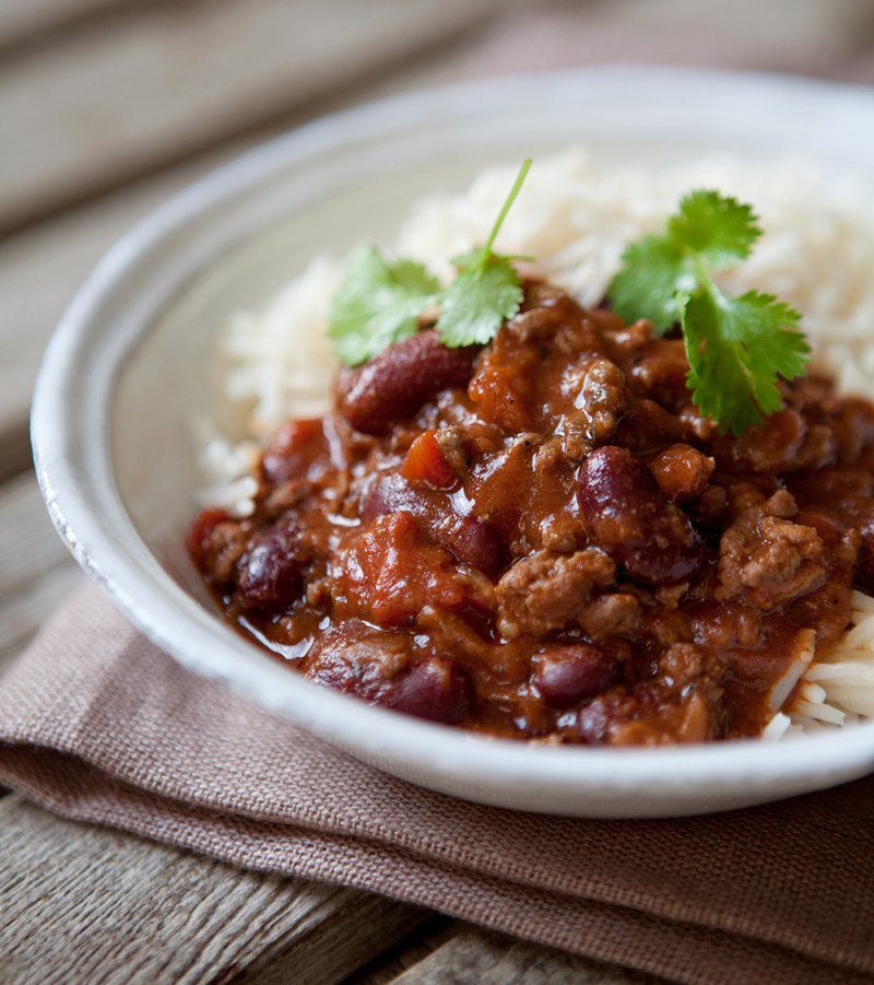 A bowl of Chilli Con Carne inspired by the Hairy Dieters Cookbook, featuring red kidney beans and minced meat over white rice, garnished with fresh coriander on a rustic wooden table.