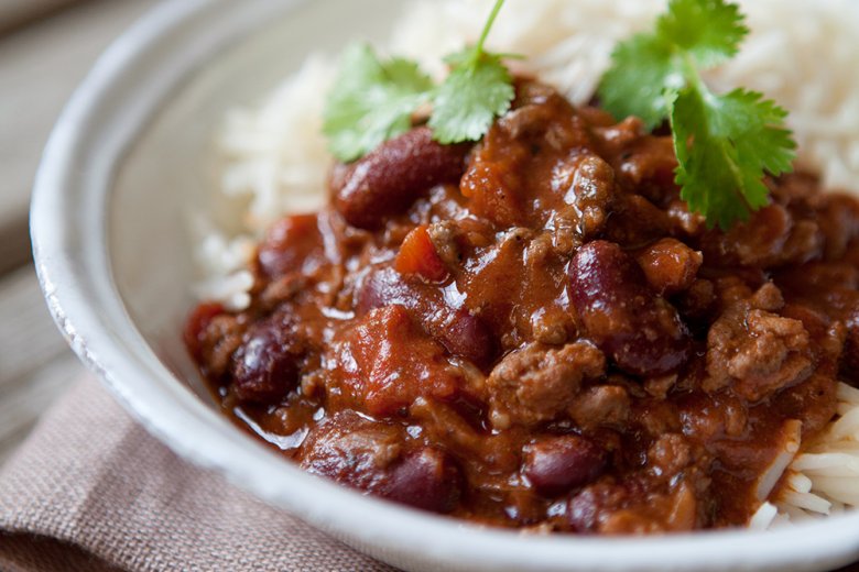 A bowl of Chilli Con Carne inspired by the Hairy Dieters Cookbook, featuring red kidney beans and minced meat over white rice, garnished with fresh coriander on a rustic wooden table.