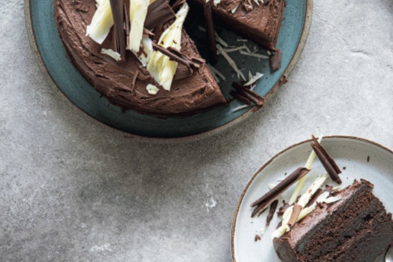 A round Chocolate Cake topped with white and dark chocolate shavings, inspired by the Hairy Bikers' Everyday Winners, with one slice served on a separate plate.