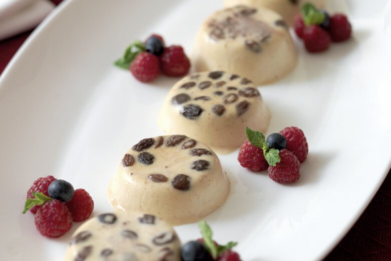 A white oval plate with five round Christmas panna cotta desserts topped with chocolate chips, garnished with fresh raspberries, blueberries, and mint leaves, set on a red tablecloth—perfect for the Hairy Bikers' 12 Days of Christmas cookery book.