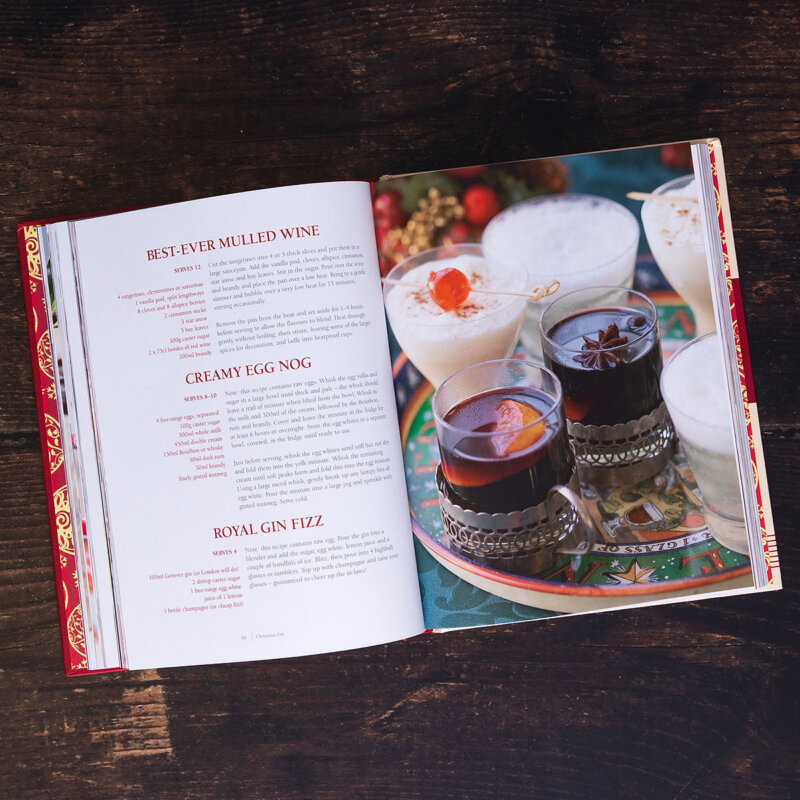 An open recipe book on a dark wooden table features 12 days of Christmas drink recipes on the left page and a festive photo, including creamy eggnog and mulled wine in glasses on a colourful tray, on the right.