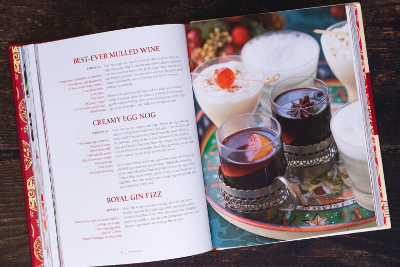 An open recipe book on a dark wooden table features 12 days of Christmas drink recipes on the left page and a festive photo, including creamy eggnog and mulled wine in glasses on a colourful tray, on the right.