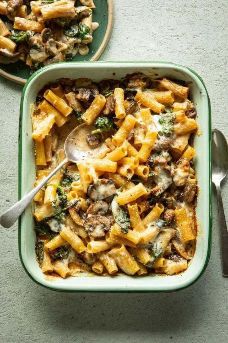 This dish is one of our family favourites: creamy pasta bake with mushrooms, spinach, and cheese in a rectangular dish, served with a silver spoon.