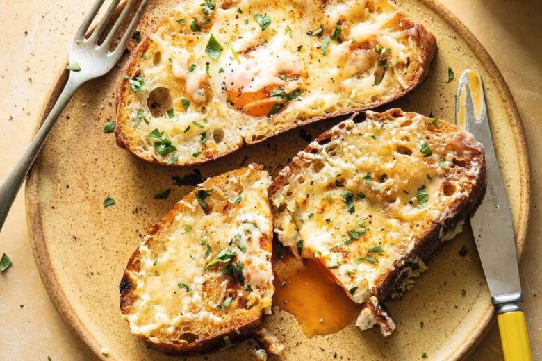 Toasted bread slices with melted cheese and herbs sit invitingly on a plate, reminiscent of family favourites. One slice is cut open, revealing egg yolk oozing perfectly, accompanied by a fork and knife—a dish the Hairy Bikers love.