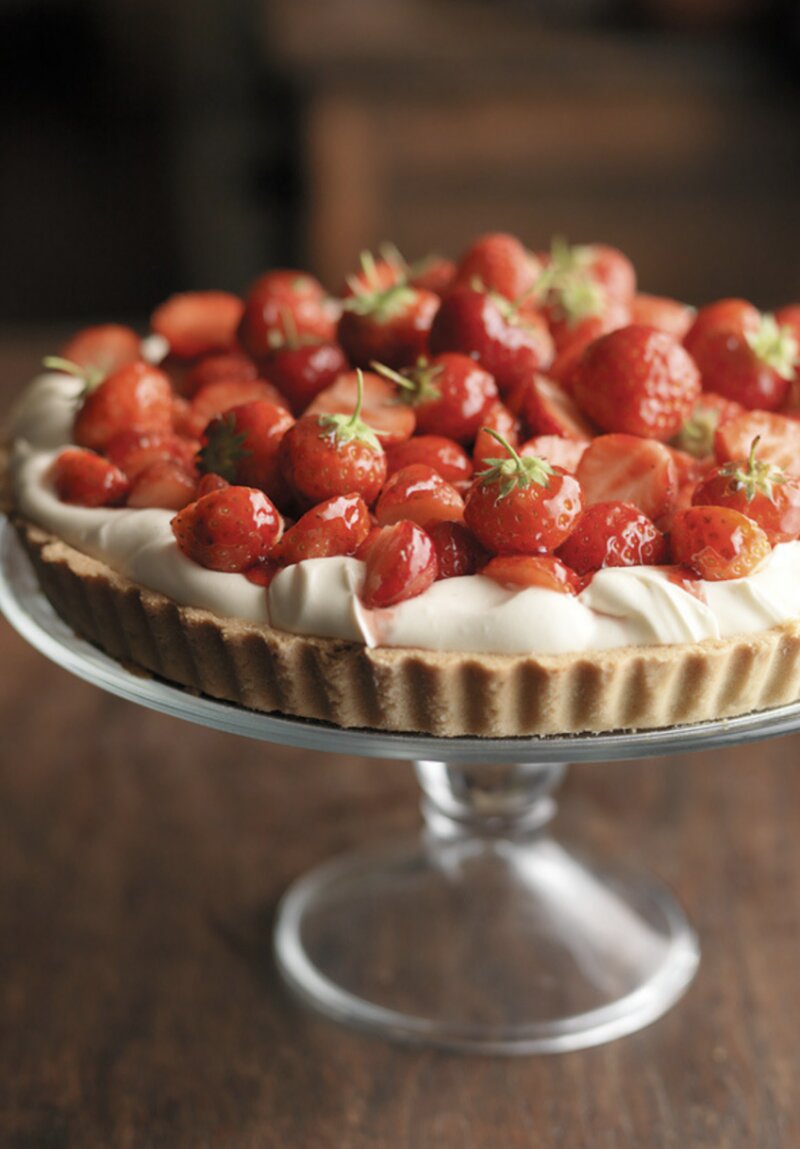 A strawberry tart topped with whole glazed strawberries sits on a glass cake stand, looking like it belongs in one of the Hairy Bikers' perfect pies recipes.