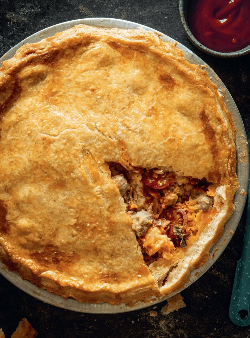 A baked pie with a slice removed, showing a filling of meat and vegetables. A small bowl of red sauce is nearby.
