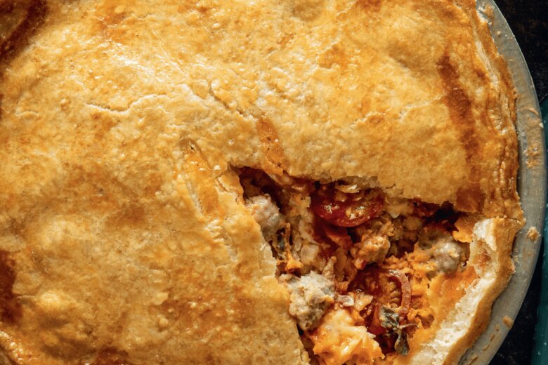 A baked pie with a slice removed, showing a filling of meat and vegetables. A small bowl of red sauce is nearby.