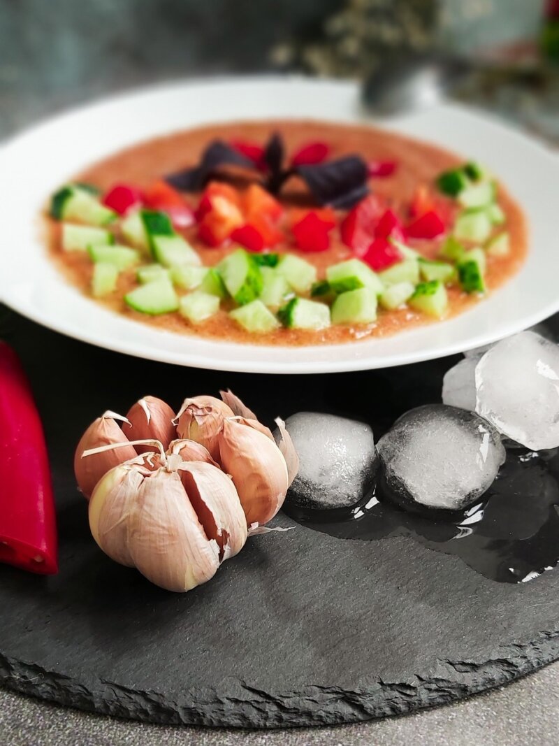 A close-up of a vegetable soup garnished with chopped cucumber and bell pepper, a cluster of garlic bulbs, ice cubes, and a red chili pepper displayed on a dark surface, reminiscent of the Hairy Bikers Everyday Gourmets' vibrant gazpacho recipe.