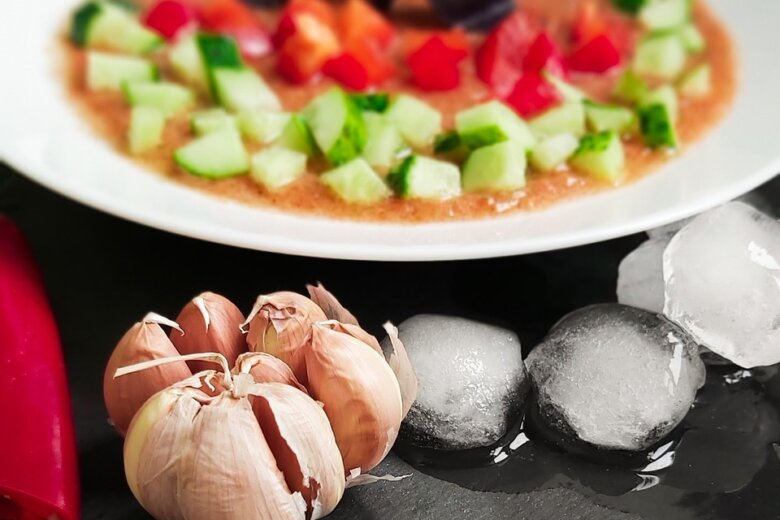 A close-up of a vegetable soup garnished with chopped cucumber and bell pepper, a cluster of garlic bulbs, ice cubes, and a red chili pepper displayed on a dark surface, reminiscent of the Hairy Bikers Everyday Gourmets' vibrant gazpacho recipe.