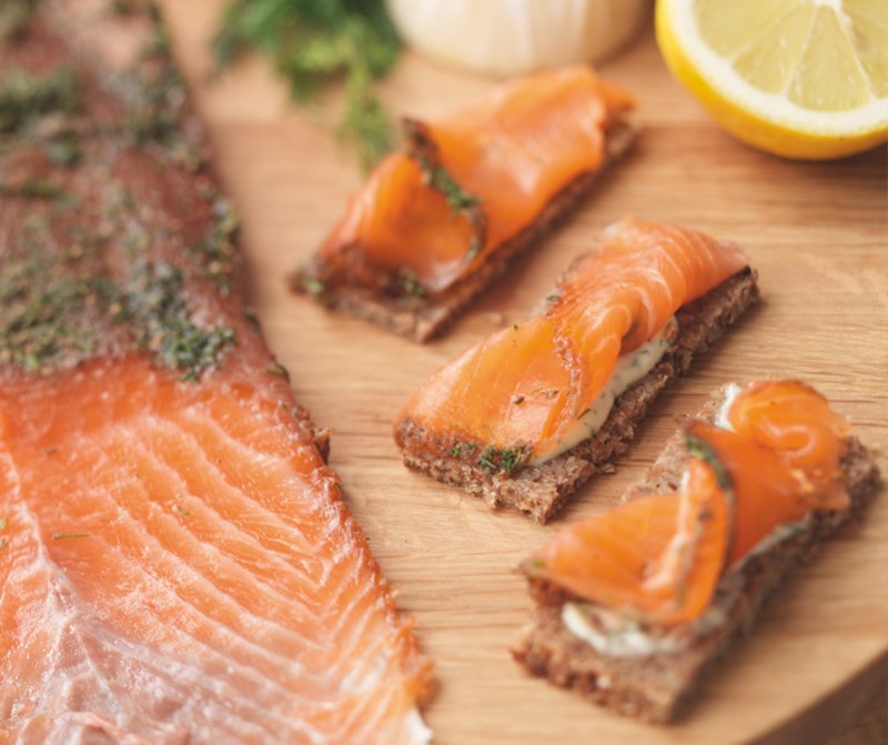 Slices of cured salmon on a wooden board, with three pieces of rye bread topped with festive Christmas gravlax and herbs. A lemon half and some herbs are visible in the background, inspired by Hairy Bikers 12 Days of Christmas.
