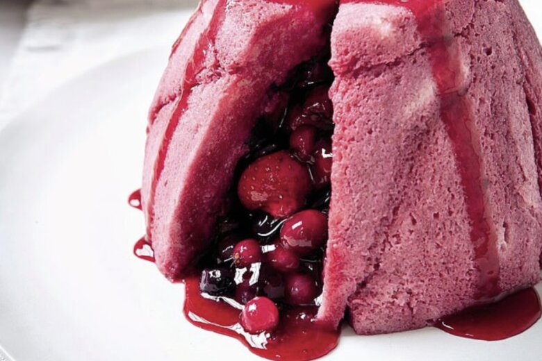A delightful summer pudding with a pink sponge base and berry filling, garnished with fresh berries on top and rich berry sauce dripping down, is placed elegantly on a white plate. Forks and plates in the background hint at good eating to come.