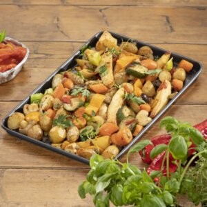 A tray of roasted vegetables including carrots, potatoes, and zucchini, garnished with herbs, served on an oven tray on a wooden table.