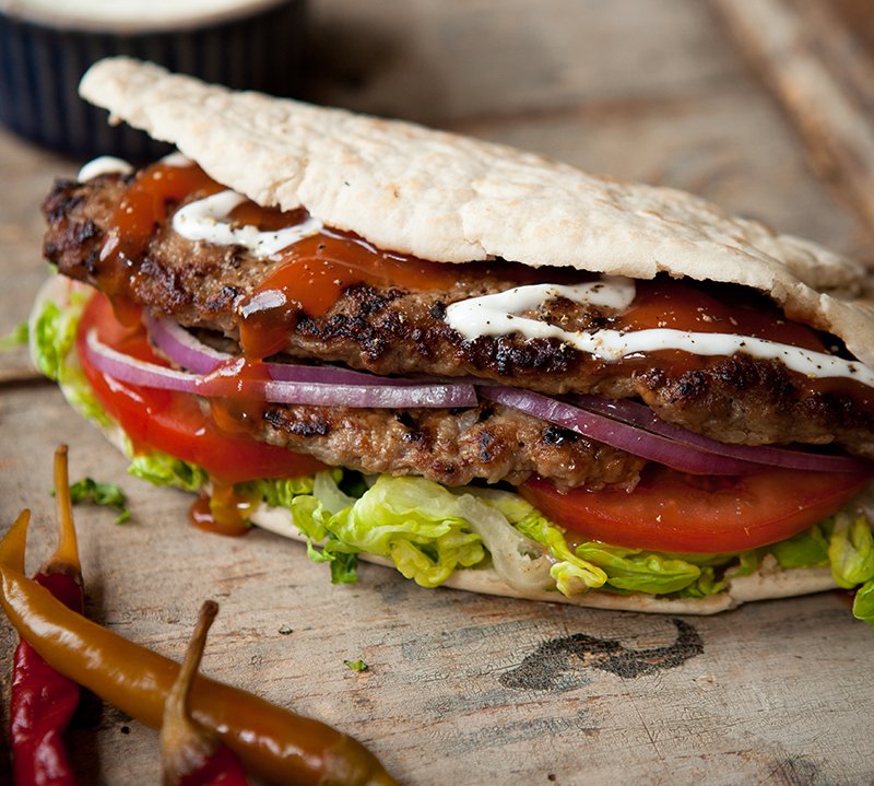 A grilled burger with lettuce, tomato, red onion, and sauces inside a folded pitta bread, reminiscent of a Hairy Bikers recipe, with chilies on the side on a wooden surface.