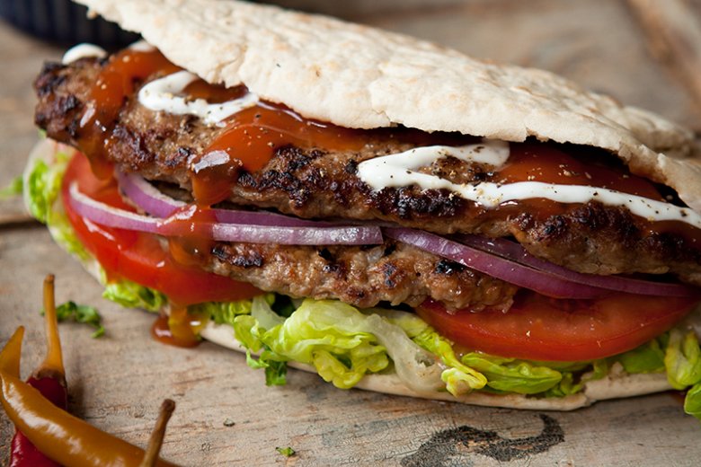 A grilled burger with lettuce, tomato, red onion, and sauces inside a folded pitta bread, reminiscent of a Hairy Bikers recipe, with chilies on the side on a wooden surface.