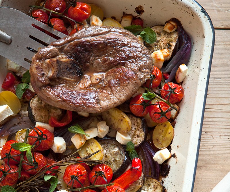 A Hairy Bikers 'Meat Feasts' favourite: a baked dish with a cooked lamb steak atop roasted vegetables—cherry tomatoes, yellow potatoes, aubergine, red onions—fresh herbs, and cubes of white cheese in a white roasting tin.
