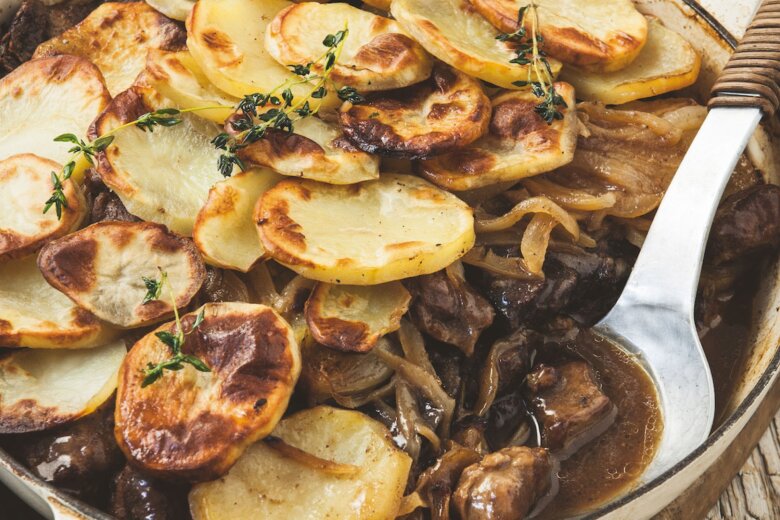 A rustic Lancashire hotpot of sliced potatoes baked atop a savoury meat and onion stew, inspired by the Hairy Bikers, served in a round ceramic pan with a serving spoon on a wooden table beside a blue serviette and glass of water.