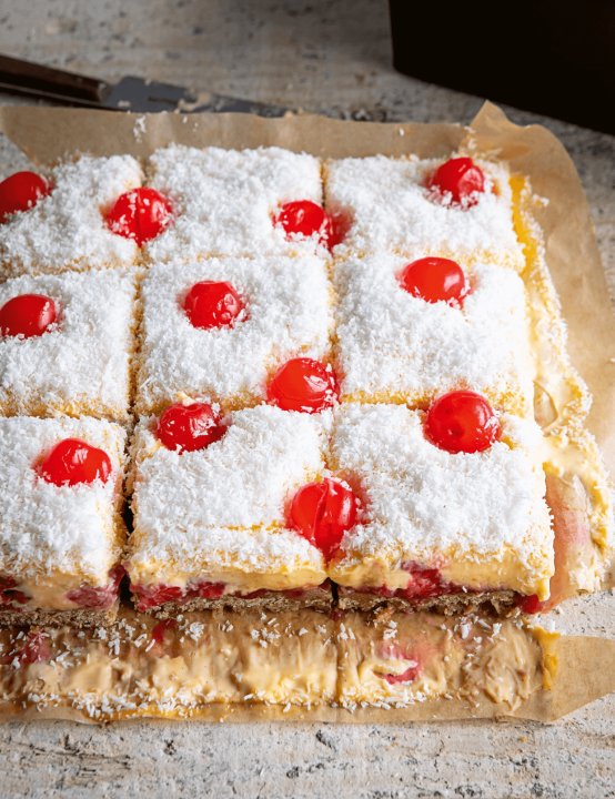 Nine square slices of Manchester Tart Flapjack, topped with shredded coconut and red cherries, are arranged on parchment paper—truly Everyday Winners for any dessert table.