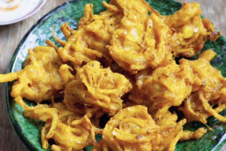 A green bowl of crispy, golden-brown pickled onion bhajis sits on a wooden table, reminiscent of a Hairy Bikers feast, with a small plate of dipping sauce in the background.
