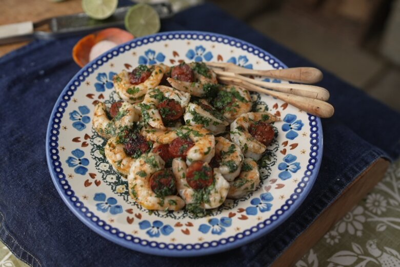 A ceramic plate with blue floral patterns showcases a shrimp dish garnished with herbs. Three wooden utensils rest on the plate's edge. The background includes lime slices, a knife, and Si King's Prawn and Chorizo bites, perfect for Simple for Summer moments.