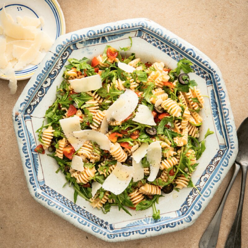 A blue and white ceramic bowl brimming with a Puttanesca Pasta Salad—pasta, arugula, cherry tomatoes, black olives, all crowned with shaved cheese. Inspired by Hairy Bikers' Fast & Fresh recipes, a small plate with extra cheese shards and silverware sits beside it.