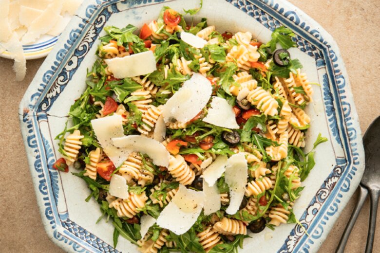 A blue and white ceramic bowl brimming with a Puttanesca Pasta Salad—pasta, arugula, cherry tomatoes, black olives, all crowned with shaved cheese. Inspired by Hairy Bikers' Fast & Fresh recipes, a small plate with extra cheese shards and silverware sits beside it.