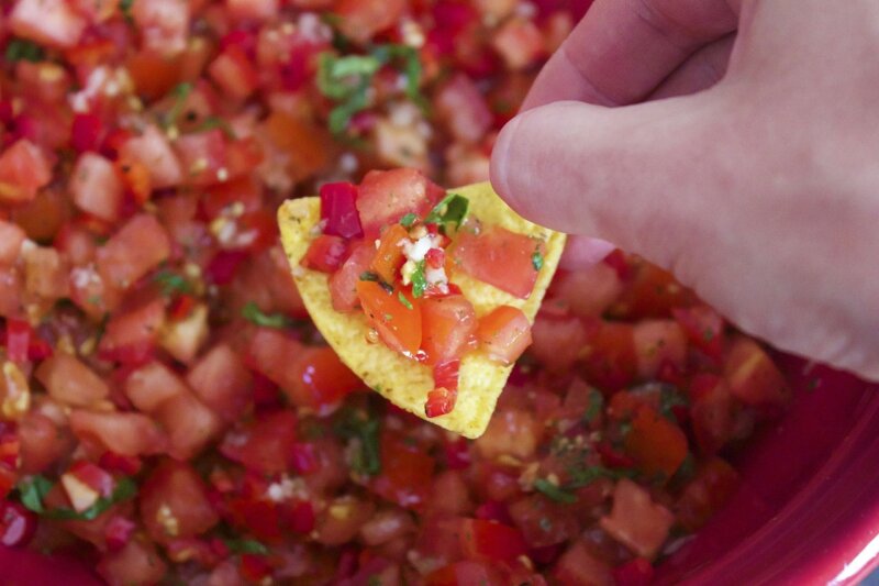 A hand holds a tortilla chip with fresh tomato salsa dip, with a bowl of the same zesty Mexican salsa in the background.