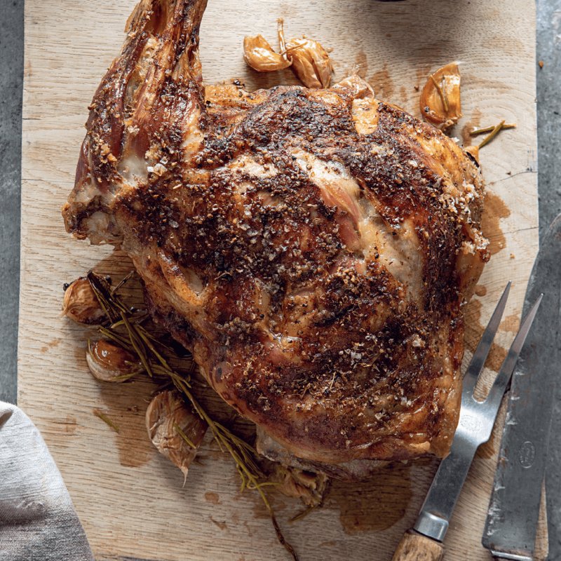 A beautifully roasted shoulder of lamb rests on a wooden cutting board, its succulent aroma mingling with garlic cloves and sprigs of rosemary. Just as the Hairy Bikers would prepare, the dish is invitingly served with a carving fork lying nearby.