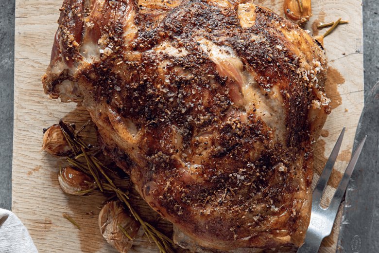 A beautifully roasted shoulder of lamb rests on a wooden cutting board, its succulent aroma mingling with garlic cloves and sprigs of rosemary. Just as the Hairy Bikers would prepare, the dish is invitingly served with a carving fork lying nearby.