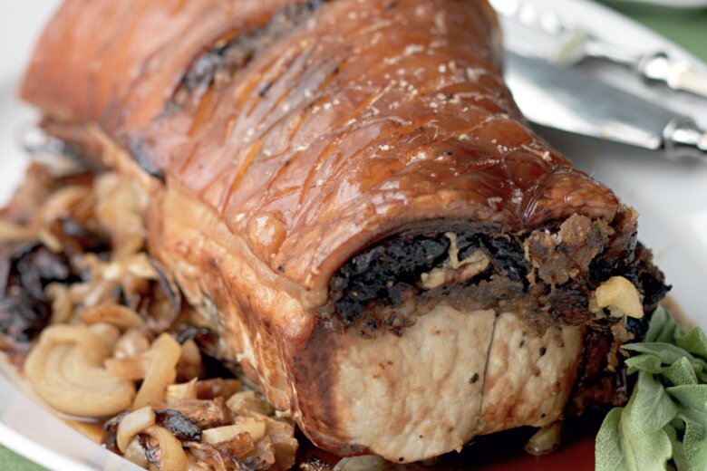 Roast Loin of Pork with crispy, golden-brown crackling rests on a plate, surrounded by caramelised onions and herbs—a classic Hairy Bikers recipe. A carving fork and knife are placed in the background.