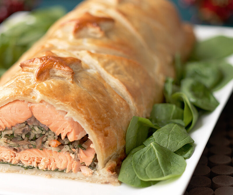 A close-up of Salmon Coulibiac, inspired by the Hairy Bikers’ Perfect Pies, with golden, flaky pastry and a pink salmon filling, sliced open and garnished with fresh spinach leaves on a white plate.