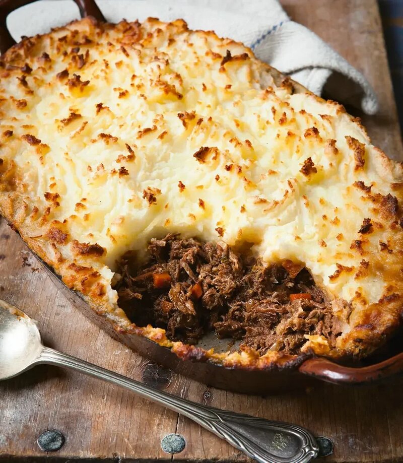 A delectable Shepherd's Pie, reminiscent of the Hairy Bikers' Best of comfort food, features a golden-brown mashed potato topping and a hearty filling of shredded beef and vegetables. A serving spoon and cloth lie ready beside this classic dish.