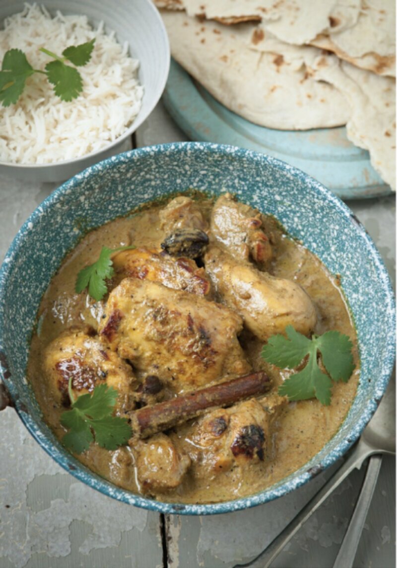 A blue bowl filled with Shorshe Murgi, a  curry garnished with coriander, accompanied by a side of white rice and flatbread in the background—a dish perfect for fans of 'Chicken & Egg cookbook by the Hairy Bikers.