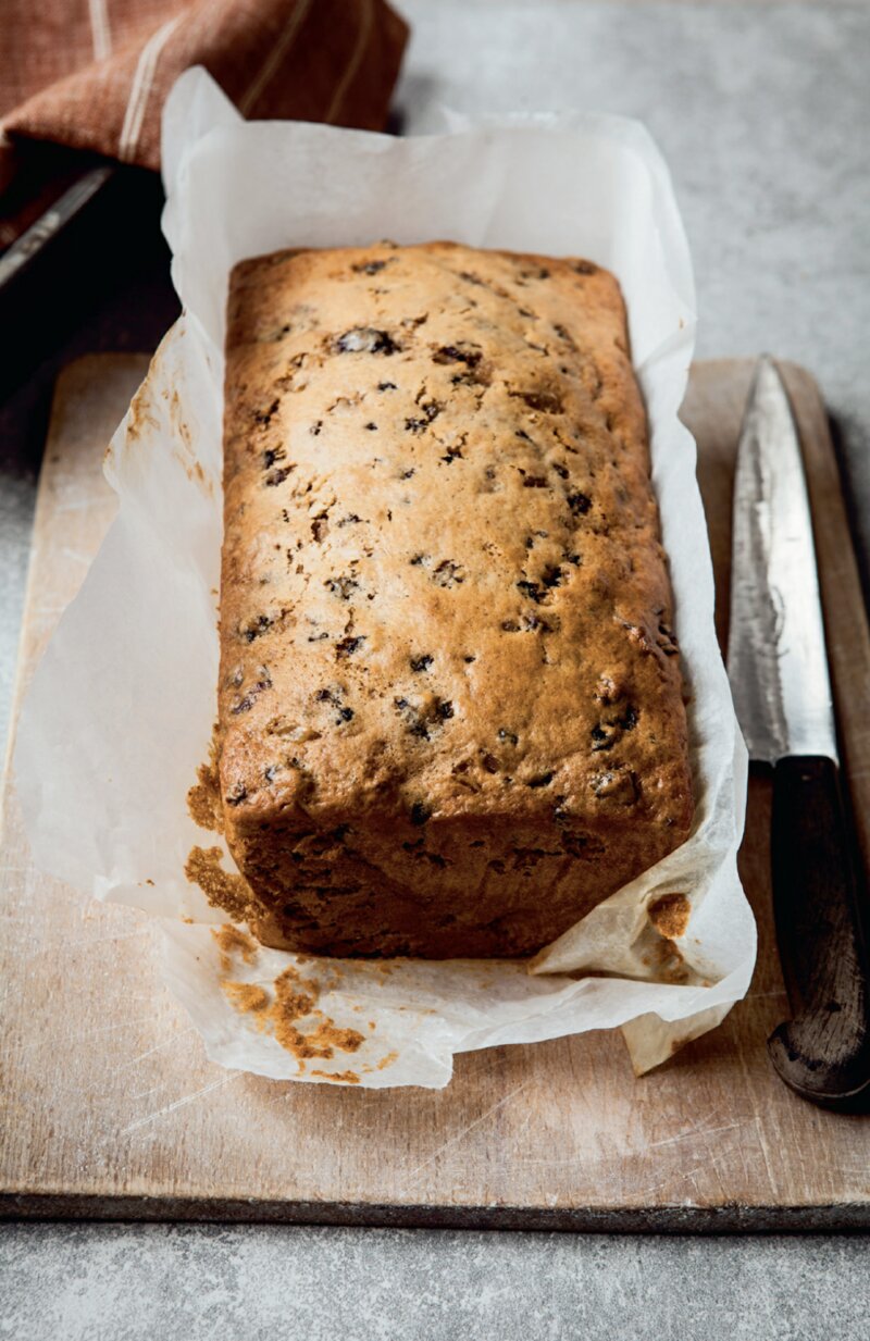 A spicy fruit loaf, from the Best of the Hairy Bikers' cookbook, rests in parchment paper on a wooden cutting board next to a bread knife.