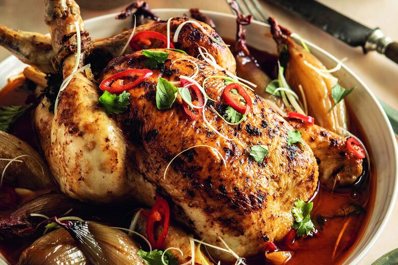 A Hairy Bikers Thai pot-roast chicken is garnished with herbs and sliced red chili, surrounded by whole onions and a rich sauce. Lime wedges rest on a small plate beside the dish, enhancing its vibrant flavours.