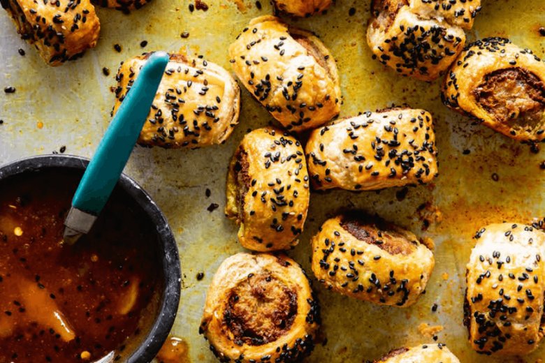 Golden, flaky Vindaloo Sausage Rolls from the Hairy Bikers Brilliant Bakes cookbook are topped with black sesame seeds and scattered on a baking tray beside a small bowl of dipping sauce with a green-handled spoon.